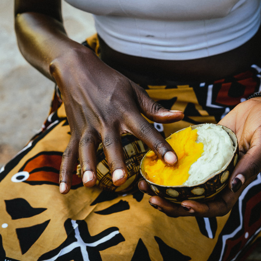 Ghanian lady in tradtitional outfit holding a wodden bowl filled with white and yellow borututu shea butter