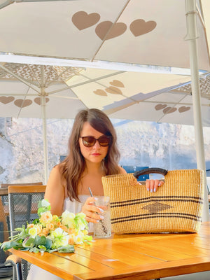 
                  
                    A woman seated at a table, sipping lemonade, casually holding a straw bag beside her, enjoying a moment of relaxation.
                  
                