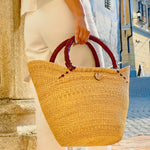 A lady holding a natural-colored, handwoven tote shopping basket with sturdy leather handles.